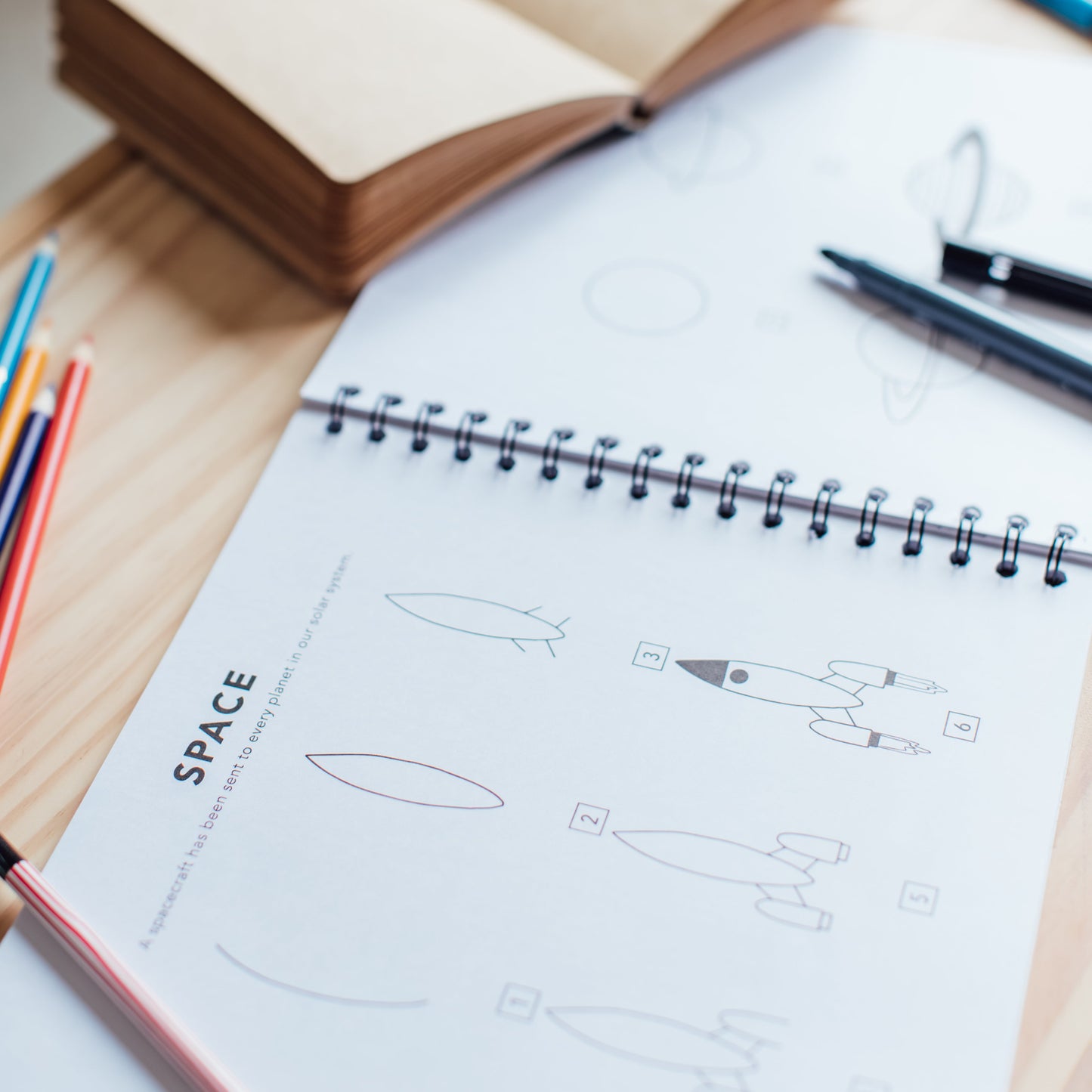 Open notebook sitting on top of "All the things: How to Draw book for Kids" showing how to draw a rocket and a planet.