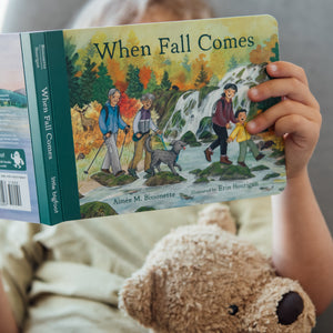 Child holding a book titled 'When Fall Comes' with a teddy bear on a couch.