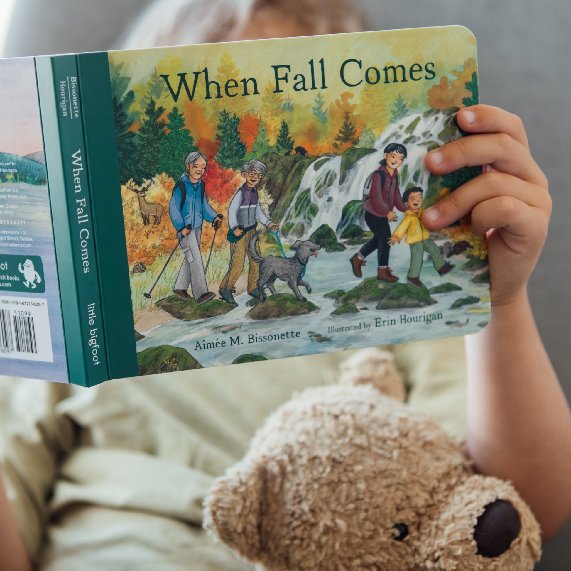 Child holding a book titled 'When Fall Comes' with a teddy bear on a couch.