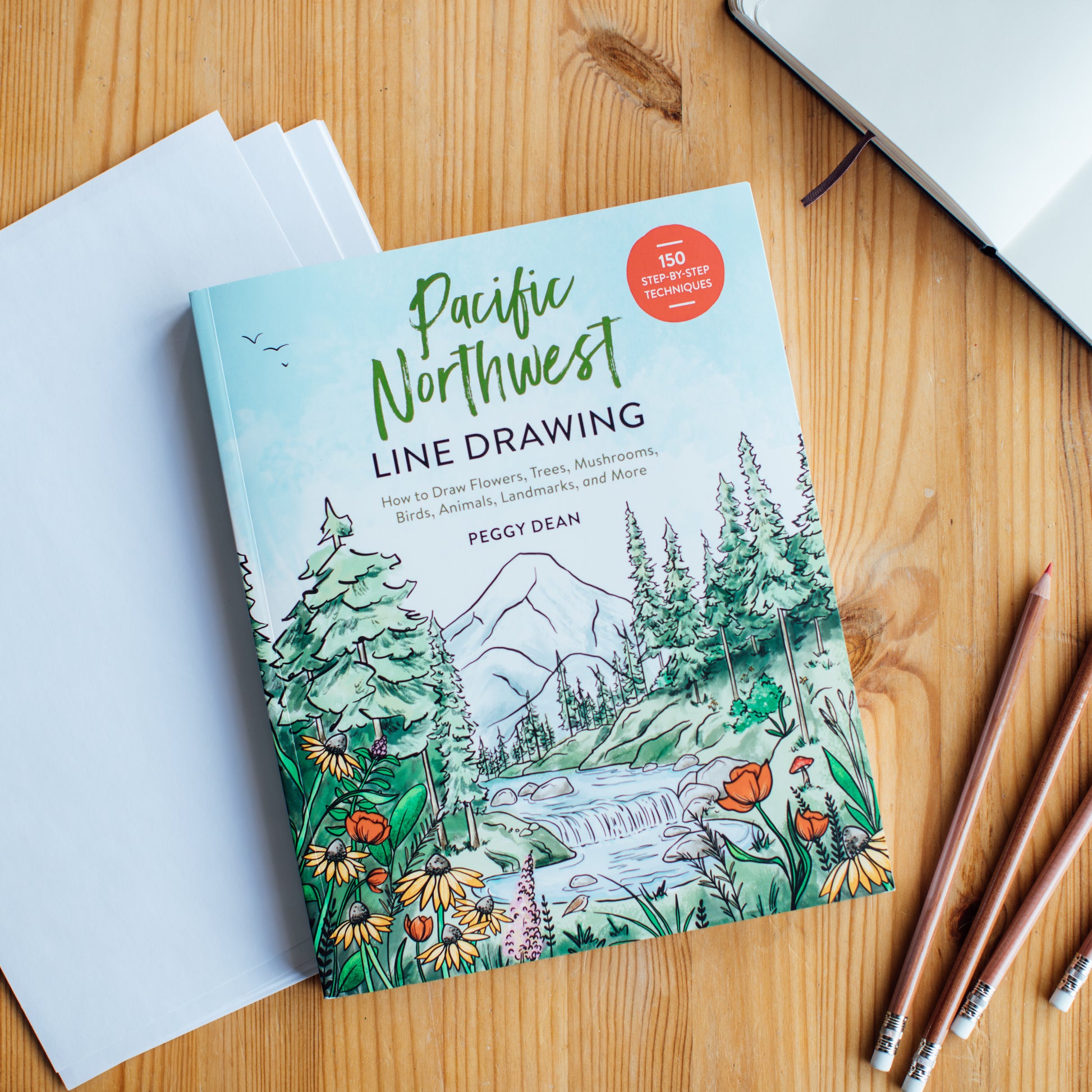 Pacific Northwest Line Drawing book with drawn images on the cover of a mountains, trees, and flowers. The Book is sitting on a wooden desk with paper and pencils. 