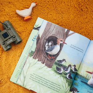 Open Book of Families of a Feather sitting on a plush orange rung with a wooden duck and some binoculars next to it. Book shares about Mergansers and how their babies are adopted. 
