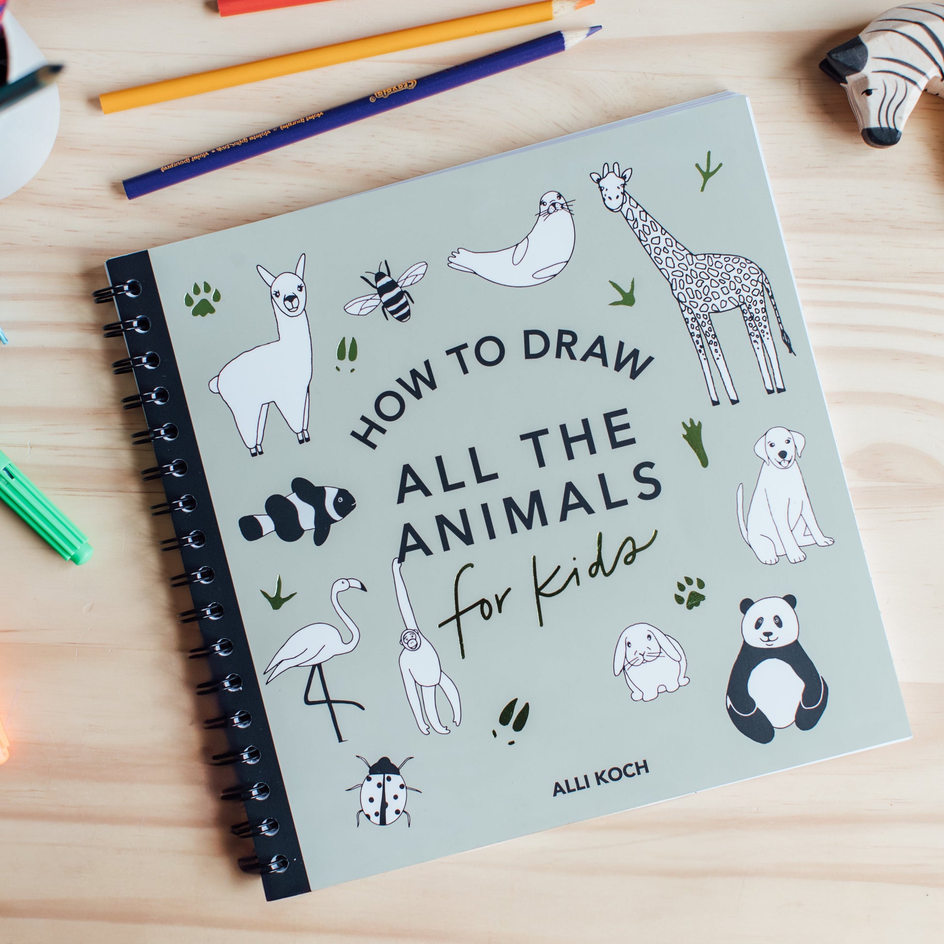 Front cover of “How to Draw All the Animals for Kids” by Alli Koch, featuring playful illustrated animals like a giraffe, panda, dog, bee, and flamingo, with a spiral binding sitting on a wooden table with colored pencils and a toy giraffe. 