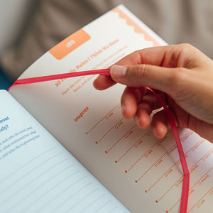 Hand holding a red string over an open book with text and a visible brand logo.
