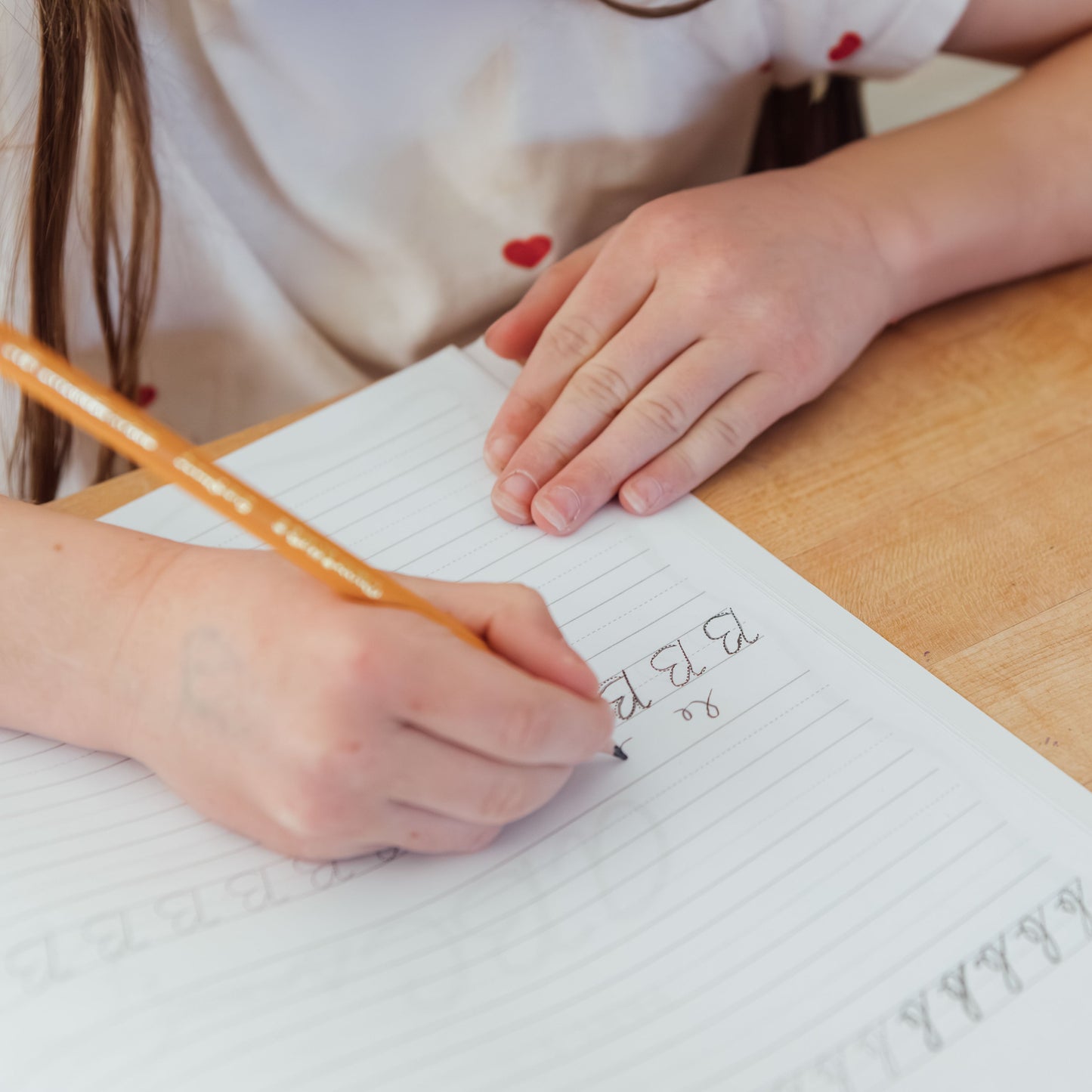 Child writing on a piece of paper with a pencil, surrounded by stationery items on a wooden table.