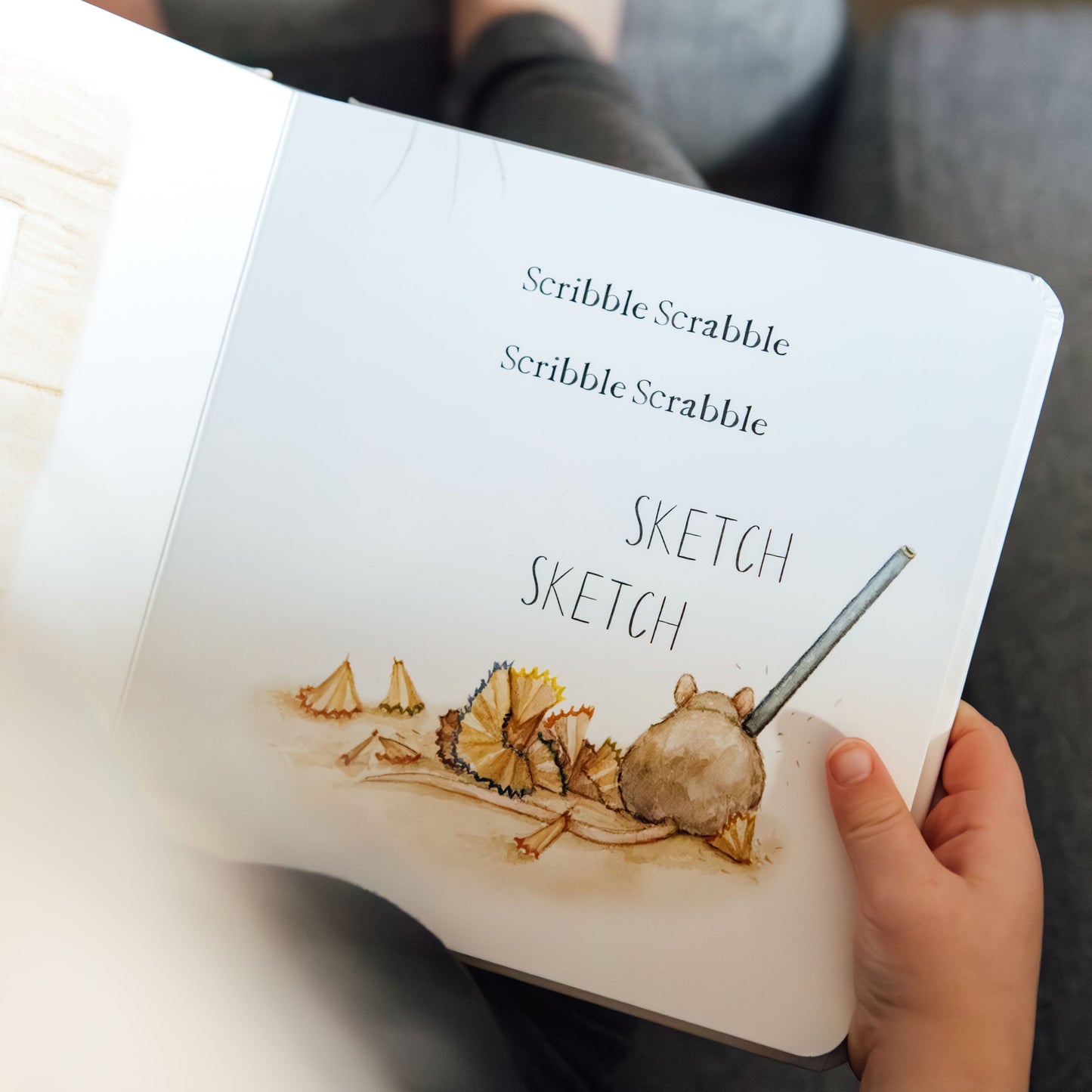 Person holding a Scribble Scrabble sketchbook with illustrations on a couch.