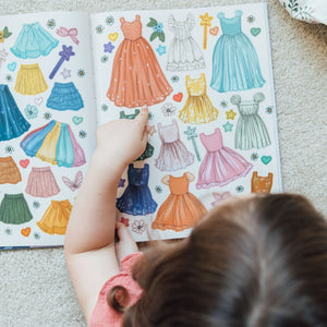 Child looking at a sticker book with colorful dress illustrations on a carpeted floor.