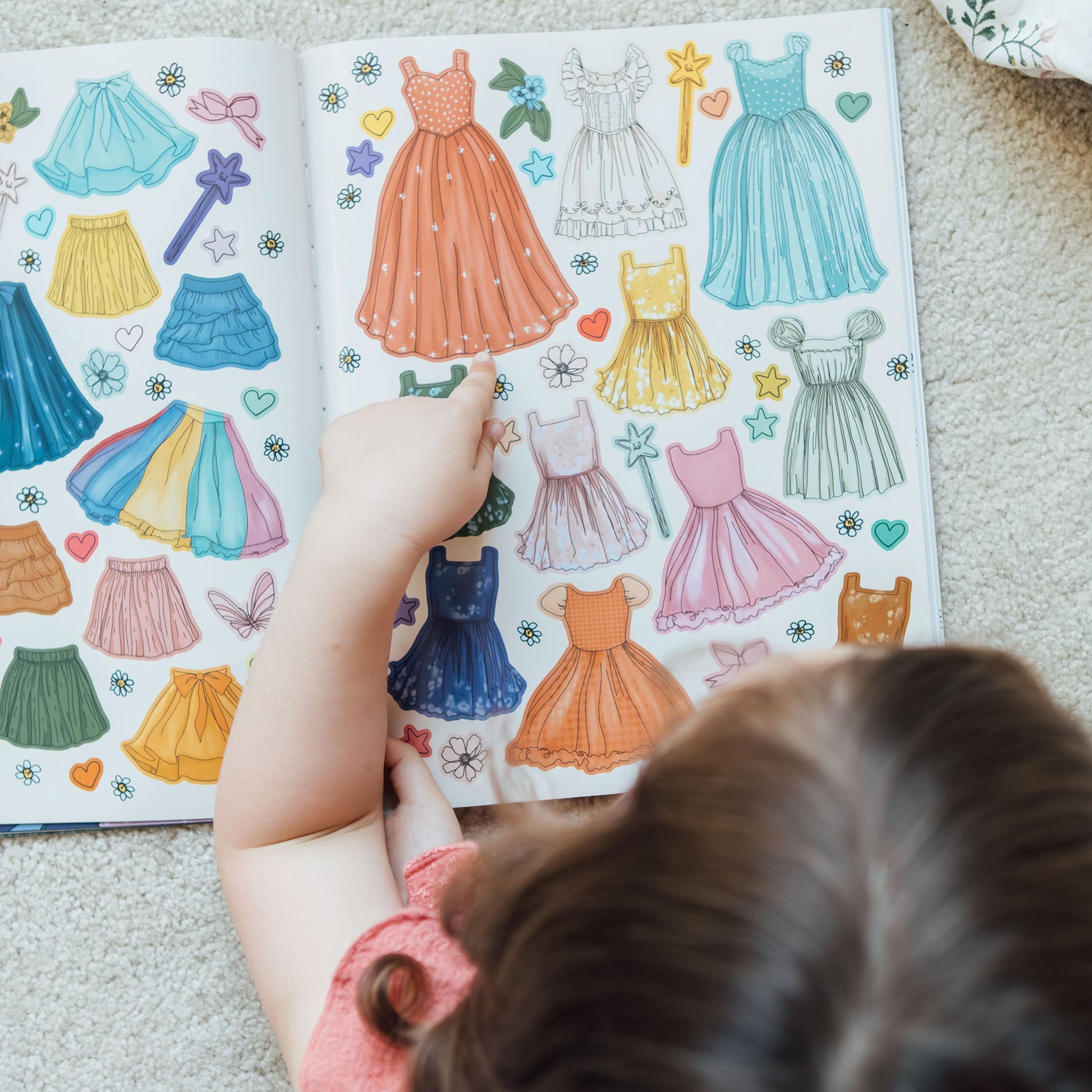 Child looking at a sticker book with colorful dress illustrations on a carpeted floor.