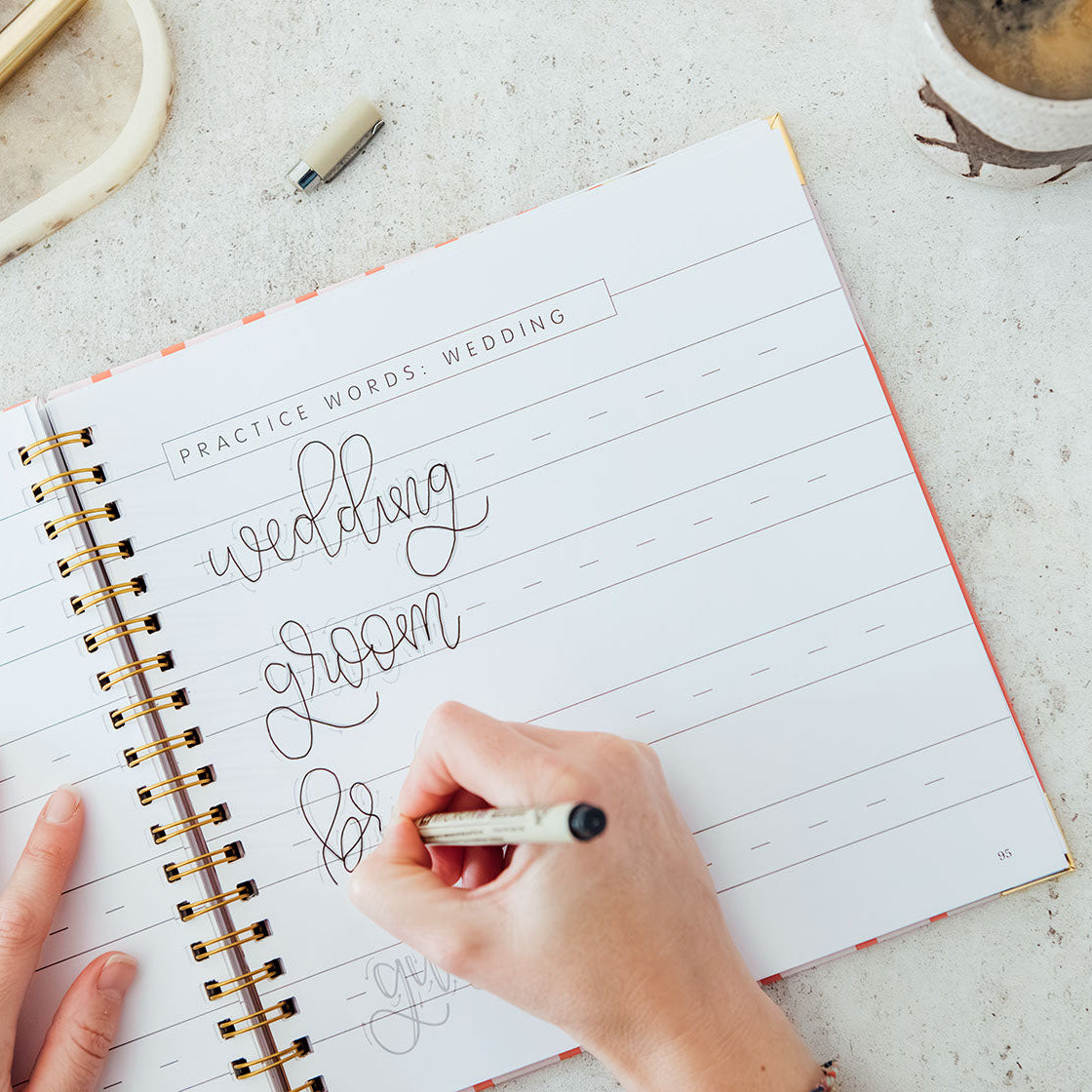 Person writing in a spiral-bound notebook with 'wedding to do list' on a light surface.