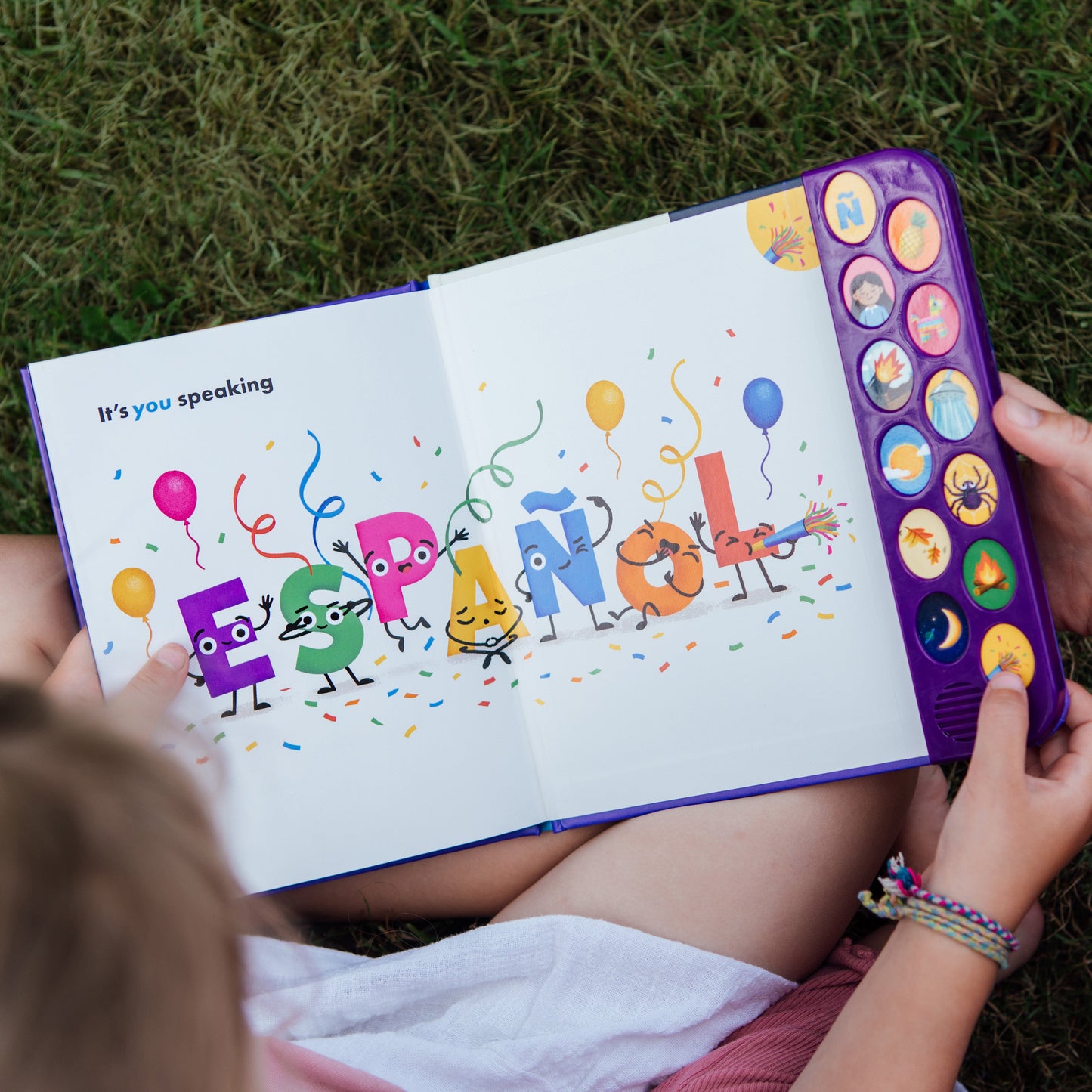 A child and an adult are sitting on grass and reading a colorful interactive children's book. The book has a purple side panel with round buttons and shows playful letter characters on the open pages.