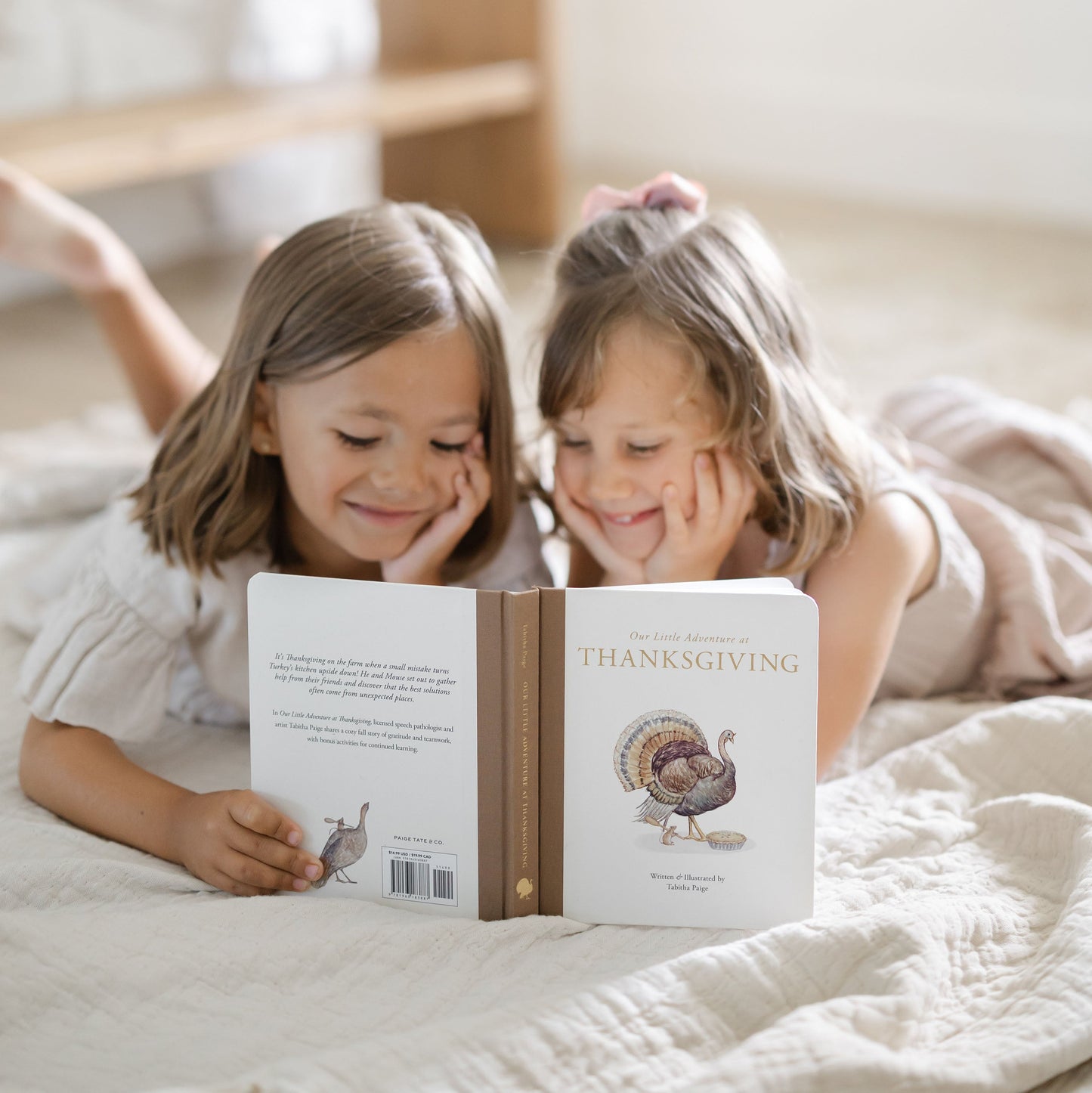 Two young girls lying on a quilted bed, smiling while looking at a personalized children's book titled 'The Little Thanksgiving' with a turkey illustration on the back cover.