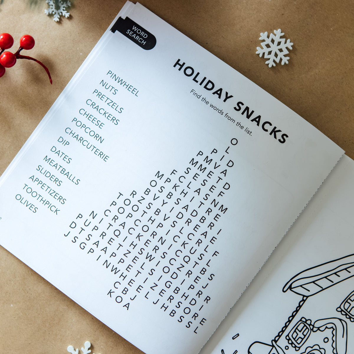 Open holiday activity book showing a “Holiday Snacks” word search puzzle shaped like a Christmas tree, with a word list including pinwheel, pretzels, charcuterie, and olives, beside a gingerbread house coloring page, styled on kraft paper with pine branches, red ornaments, berries, and white snowflakes.