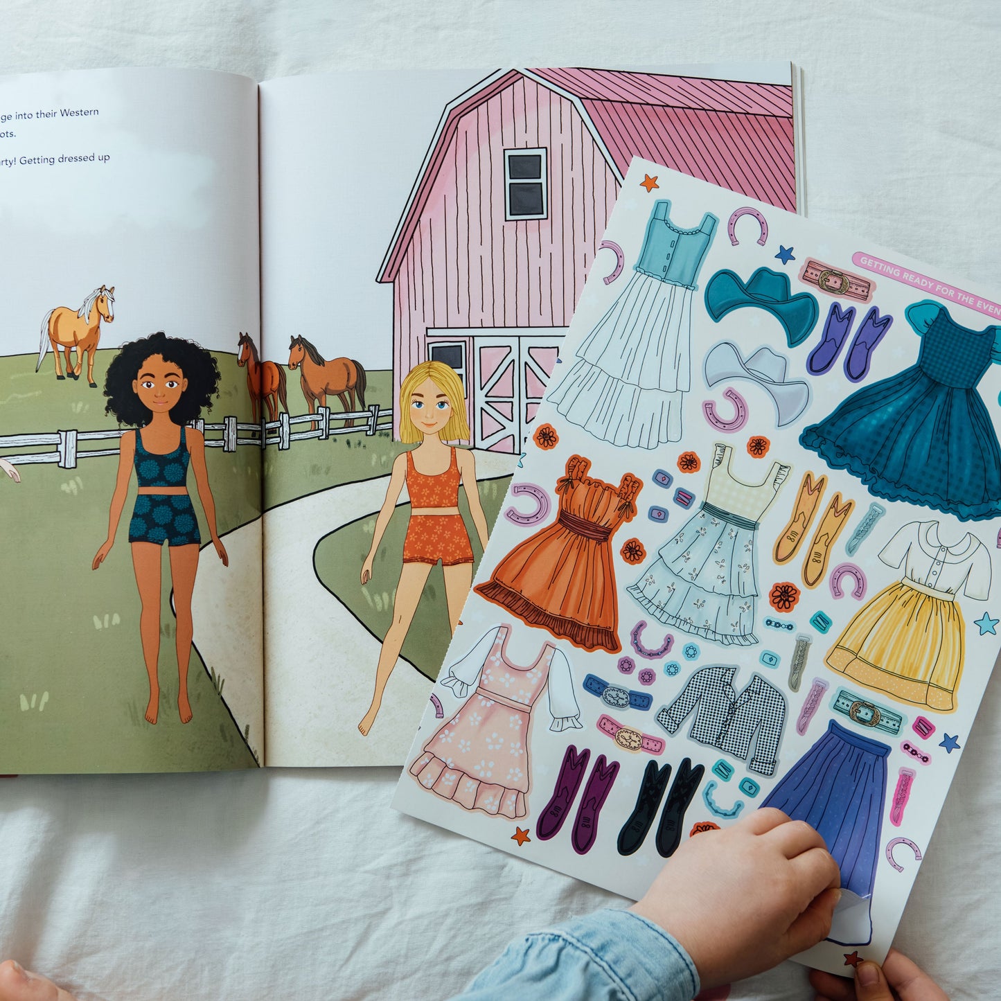 An open children's picture book with farm-themed illustrations is laid on white fabric, alongside a matching sticker sheet featuring colorful dresses, boots, and accessories. A child's hand is visible holding the sticker sheet.