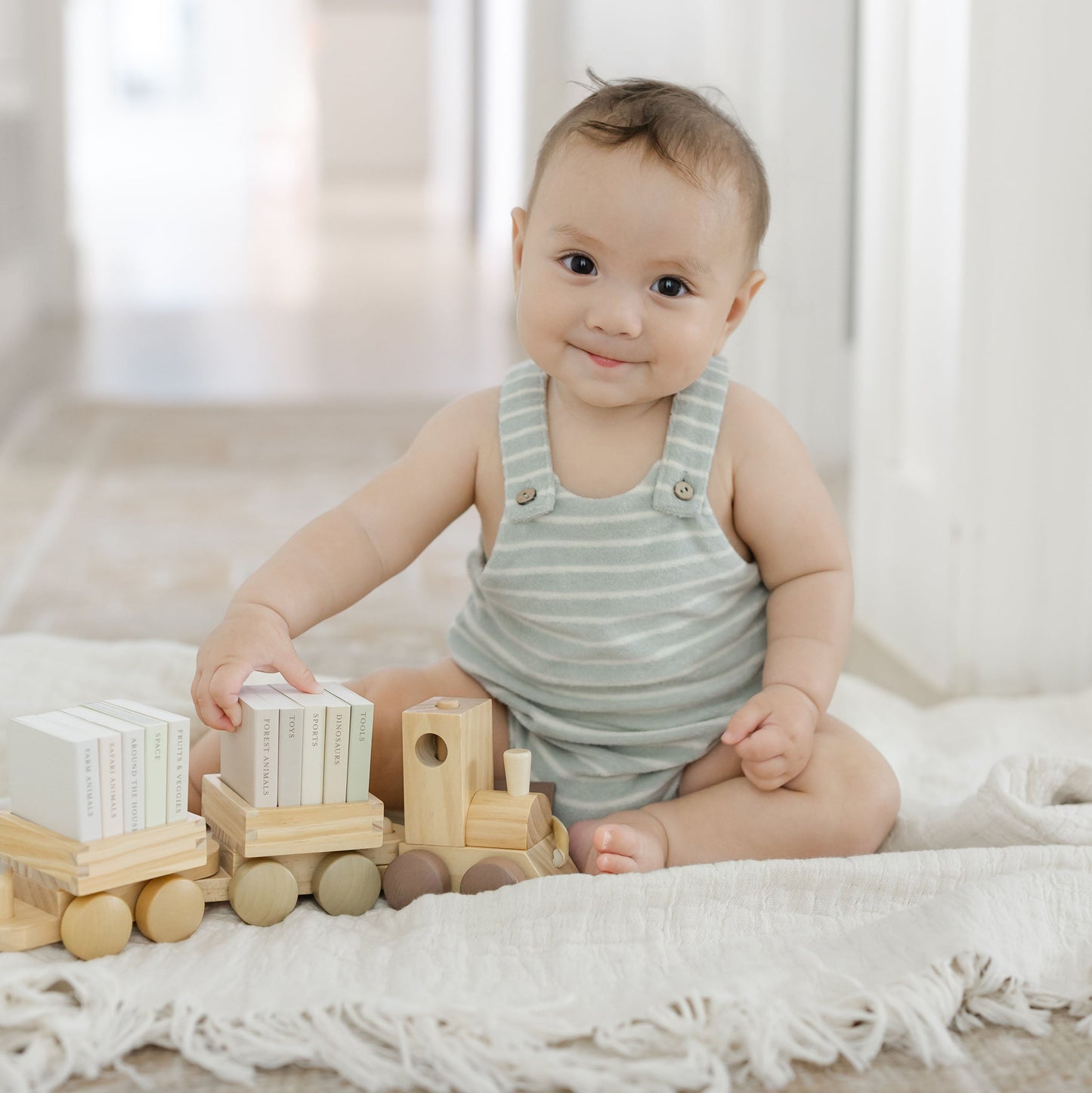 A baby sitting on a white fringed blanket playing with a wooden toy train set in a bright, modern room.