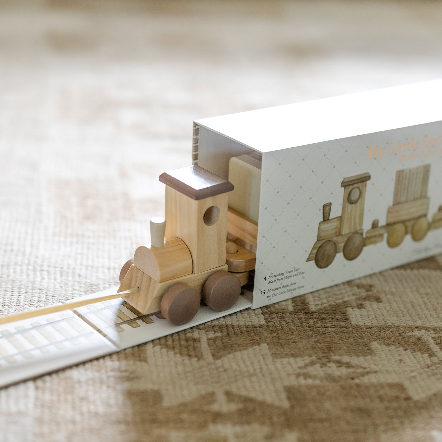 A wooden toy train set in its white packaging box, photographed on a textured beige fabric surface. The box features an illustration of the train and the text 'My Little Expression Livery'.