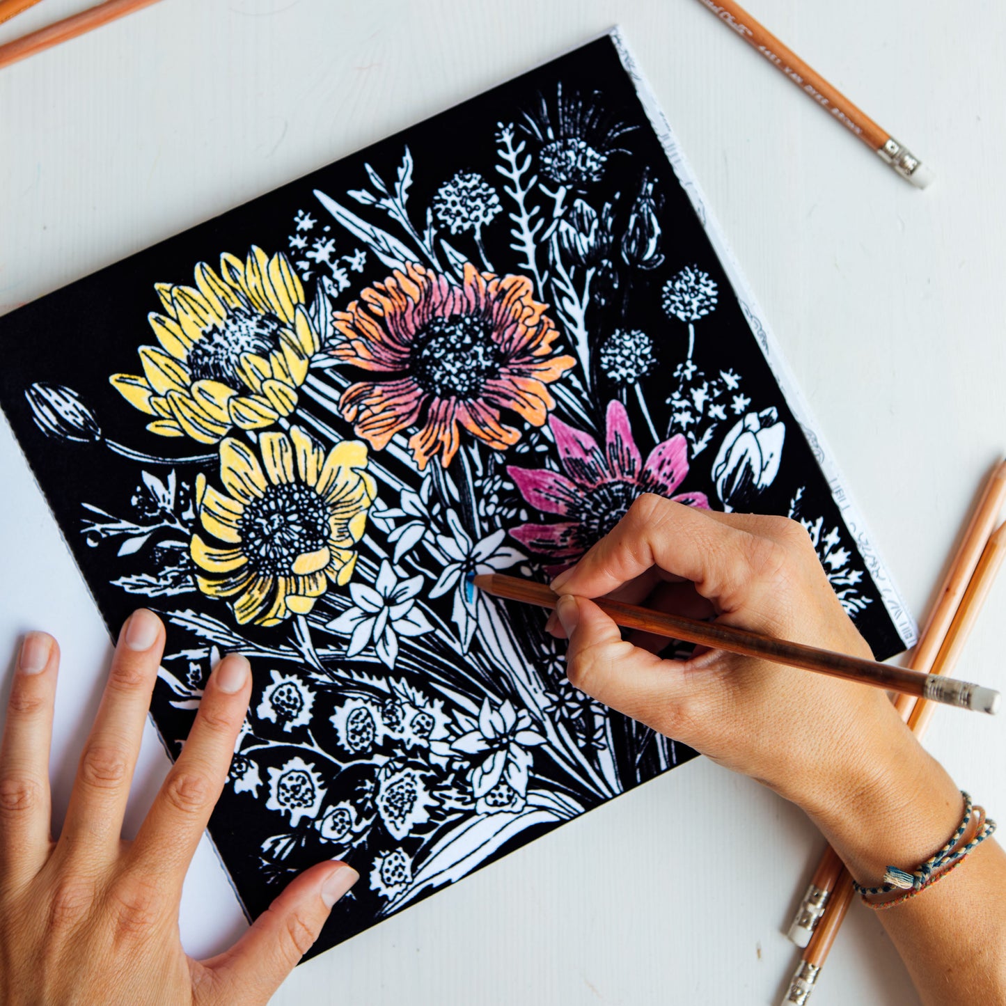 A top-down view of a person's hands coloring a black canvas with floral linocut or stenciled designs. The canvas features yellow, orange, and pink flowers with white outlines.