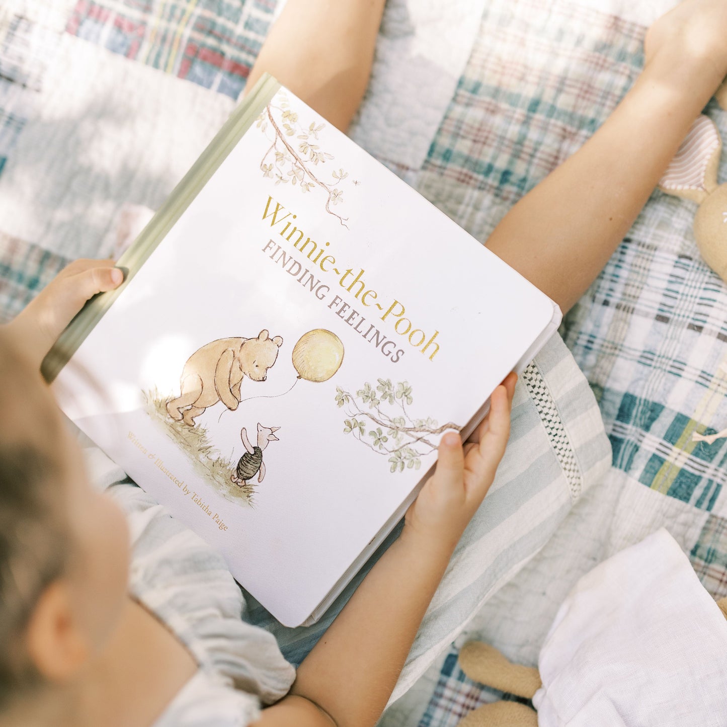 A child holds a large illustrated book titled "Winnie-the-Pooh Finding Feelings" while sitting on a plaid blanket. The book cover shows Winnie-the-Pooh and a rabbit with a yellow balloon, and the photo is taken in a soft, sunlit indoor setting.