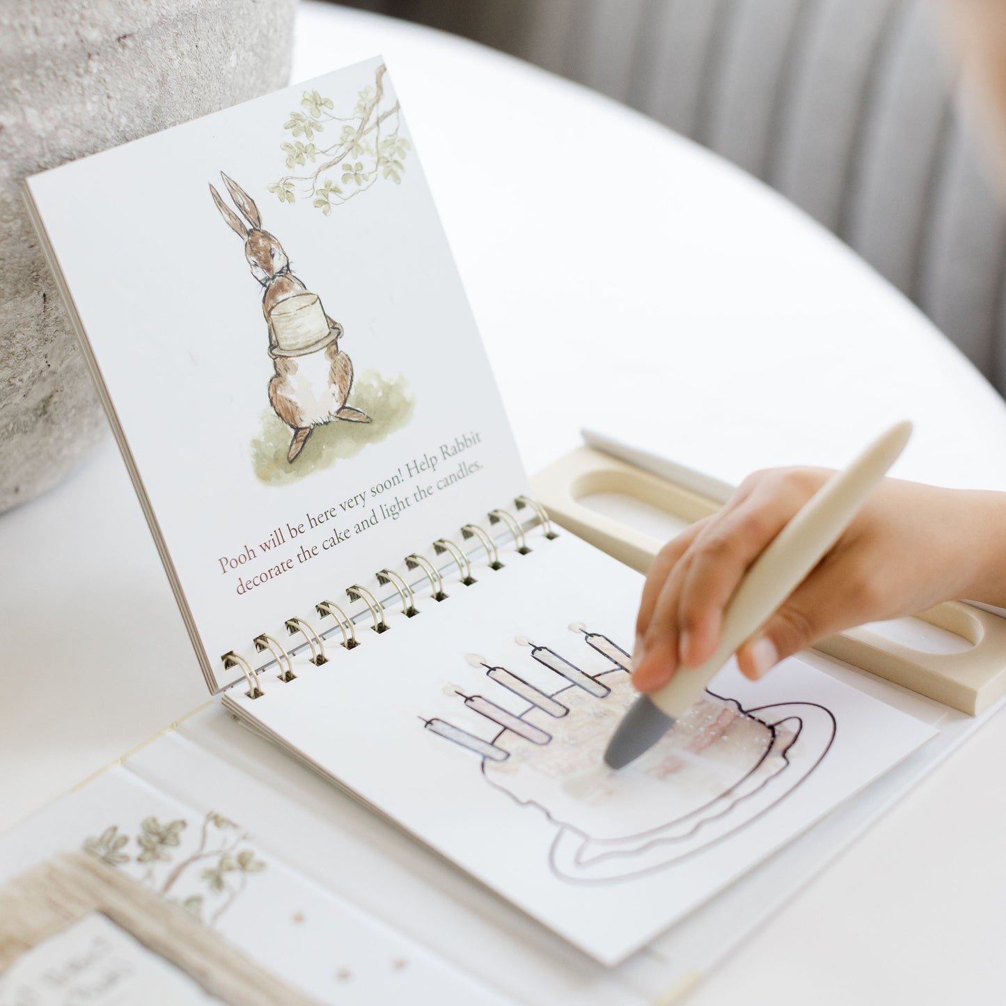 A child's hand holding a wooden paintbrush and coloring a page in a spiral-bound children's activity book. The book features a watercolor rabbit illustration on the cover and is placed on a white table next to a textured gray vase.
