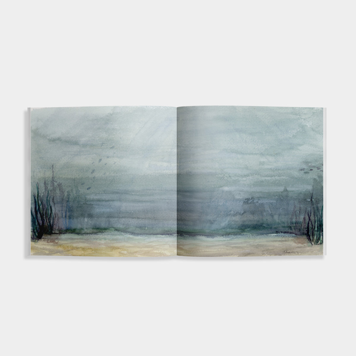 An open watercolor landscape art print showing a misty, atmospheric seascape with muted blue-gray tones, dark reeds, and a pale sandy shore, photographed against a light gray background.