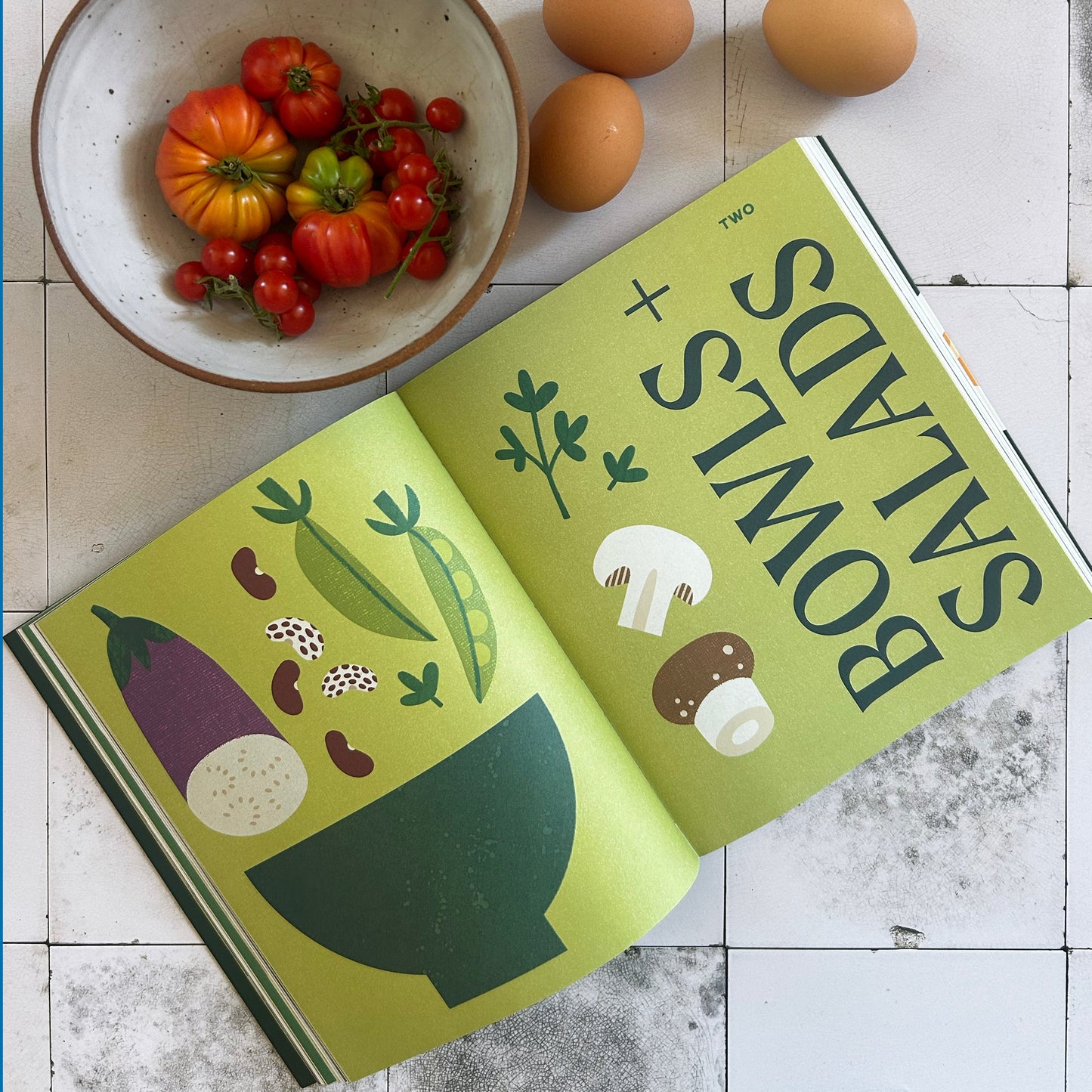 Open book titled 'Bowls + Salads' with a bowl of tomatoes and eggs on a tiled surface